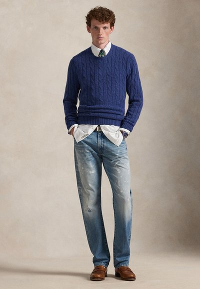 Noor mees, kes kannab sinist kaabelmustriga kampsunit, valget särki rohelist triibulist lipsu, pleekinud teksapükse ja pruune nahast kingi, seisab käed taskutes.