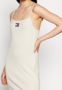 Béžové žebrované šaty s tenkými ramínky, zdobené malou polepkou v červené a modré barvě. Přiléhavý střih s bočními výřezy, hladká textura.