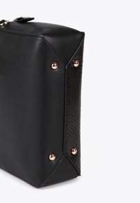 Angolo di una borsa in pelle nera che mostra il lato texturizzato, con dettagli in hardware dorato e borchie, su uno sfondo bianco semplice.