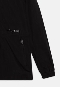 Svart jacka med en stilren design, blixtlåsficka i sidan, elastiska ärmslut och subtil logotypdetalj på framsidan. Slät textur.