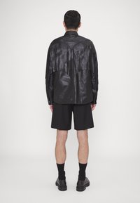 Camisa de cuero negra con flecos, mangas largas y un diseño de yugo curvado, combinada con pantalones cortos negros y calzado negro.