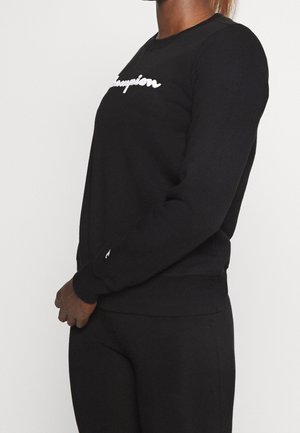 Persoon draagt een zwart sweatshirt en broek met wit "Champion" logo op de borst, staand in zijaanzicht tegen een effen achtergrond.