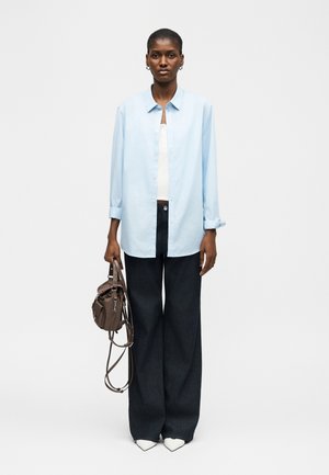 Femeie purtând o cămașă bleu cu mâneci lungi, top alb, pantaloni largi negri, pantofi albi ascuțiți, ținând o geantă maro pe lângă corp pe fundal alb.