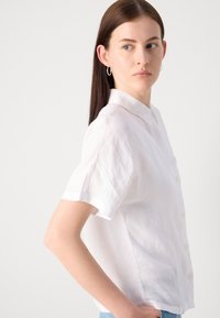 Camisa de linho branca com botões, mangas curtas, colarinho e um corte descontraído. Apresenta botões amarelos e um tecido macio e texturizado.