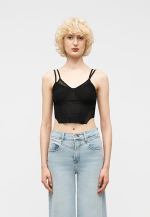 Mladá žena s krátkymi kučeravými blond vlasmi, oblečená v čiernom priesvitnom krátkom tielku a svetlomodrých džínsoch s vysokým pásom, stojaca vzpriamene.