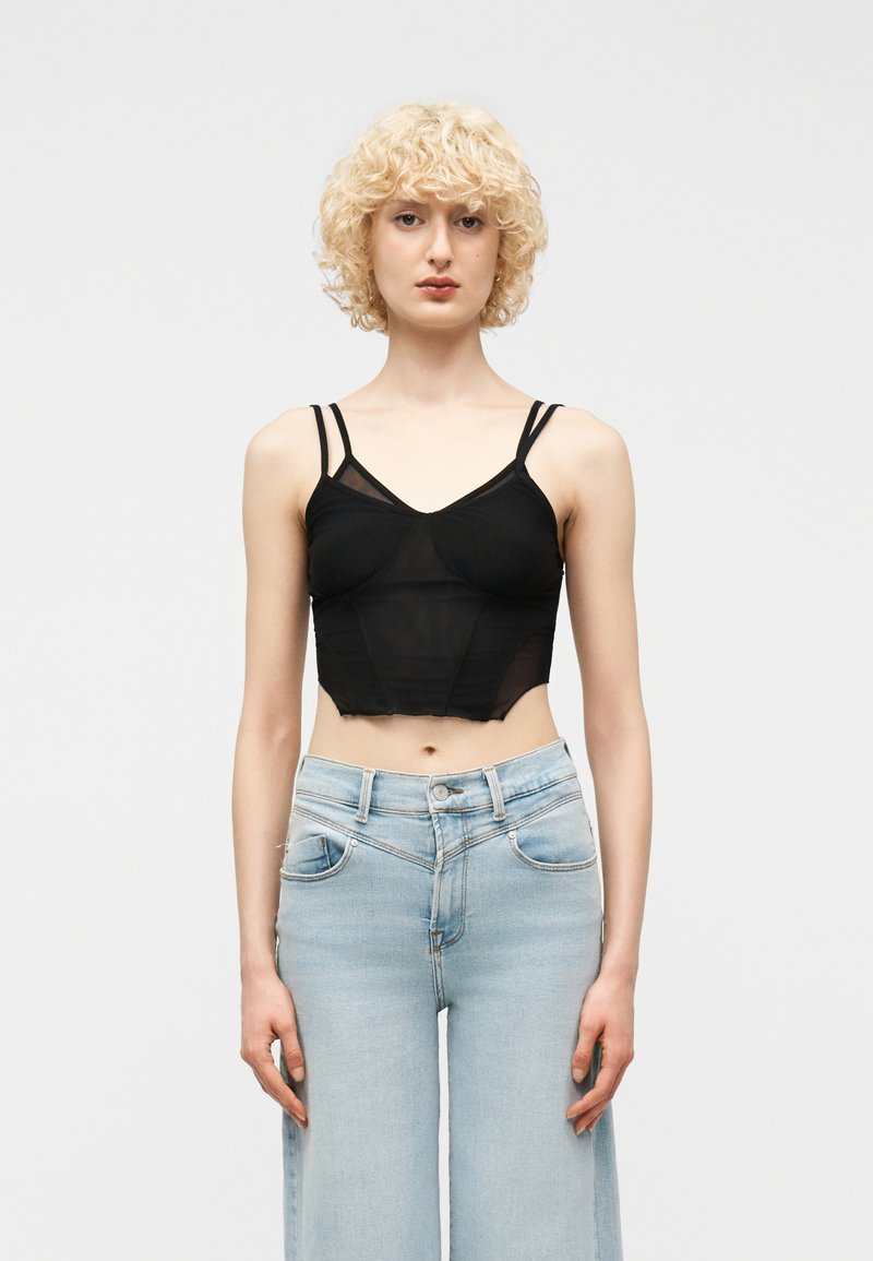 Νέα γυναίκα με κοντά, σγουρά ξανθά μαλλιά που φοράει μαύρο διάφανο κορμάκι και ανοιχτόχρωμα, ψηλόμεσα τζιν, στέκεται όρθια.