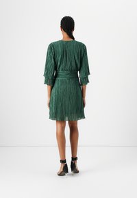 Grøn plisseret kjole med en tætsiddende talje, trekvart pufærmer og en svævende kant. Stoffet har en tekstureret glans. Sorte strophæle.