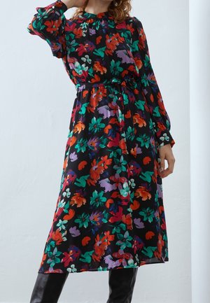 Femme portant une longue robe noire avec des motifs floraux colorés rouges, verts, violets et oranges et des bottes noires, debout contre un mur blanc.