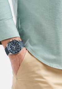 Orologio Nautica con cinturino in silicone blu e cassa in acciaio inossidabile, con quadrante blu e dettagli argentati, indossato con una camicia verde menta e pantaloni khaki.