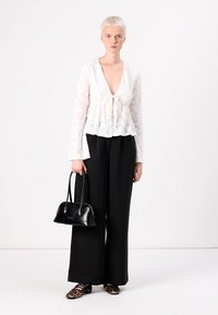Weiße Spitzenbluse mit V-Ausschnitt und geraffter Taille, kombiniert mit schwarzen weiten Hosen. Das Model hält eine schwarze Handtasche und trägt Schuhe mit Leopardenmuster.