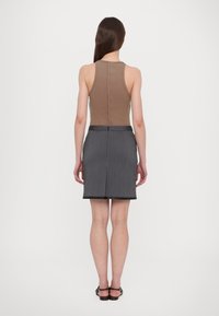 Donna con lunghi capelli castani che indossa una canotta marrone senza maniche, una gonna grigia a righe sottili e sandali neri scoperti sul retro, in piedi su uno sfondo bianco.