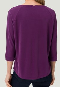 Frau, die ein lila Langarmshirt mit einem kleinen Schlitz im hinteren Halsausschnitt trägt, kombiniert mit dunkelblauen Jeans, von hinten gezeigt.