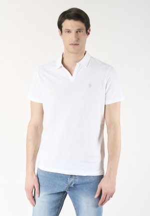 Uomo che indossa una semplice polo bianca a maniche corte e jeans azzurri, in piedi su uno sfondo neutro.