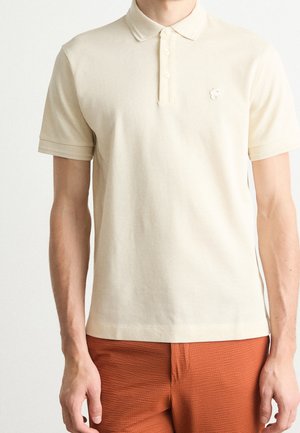 Mand iført en kortærmet, offwhite polo med et lille broderet emblem og teksturerede orange bukser.