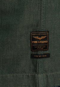 Étiquette en tissu vert en chevrons avec des accents noirs et dorés ; texte « PME LEGEND » et « TESTÉ À HAUTE ALTITUDE », étiquette de taille en bas.