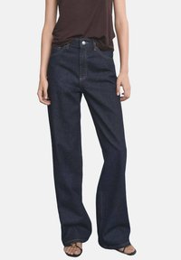 Jean en denim bleu foncé évasé avec poches avant, braguette zippée et surpiqûres contrastées. Le tissu semble robuste avec une texture lisse.