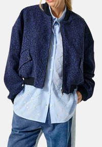 Chaqueta bomber azul marino en tejido texturizado, con cierre de cremallera en la parte delantera, puños acanalados y un bolsillo lateral. Llevada sobre una camisa de patrón azul claro.