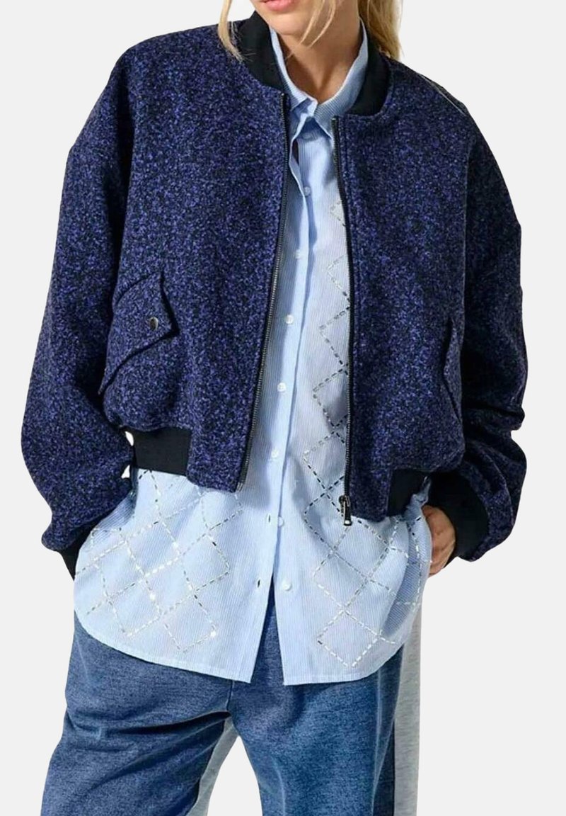 Chaqueta bomber azul marino en tejido texturizado, con cierre de cremallera en la parte delantera, puños acanalados y un bolsillo lateral. Llevada sobre una camisa de patrón azul claro.