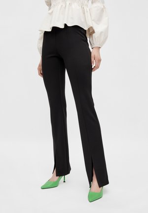 Zwarte op maat gemaakte broek met zijsplits en een gladde textuur, gecombineerd met groene puntige schoenen, gedragen met een crèmekleurige top met ruches.