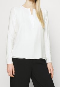 Biała bluzka z długim rękawem i delikatnym dekoltem w kształcie litery V, o miękkiej fakturze i prostym wzorze, zestawiona z czarnymi spodniami. Minimalistyczny styl i czyste linie.