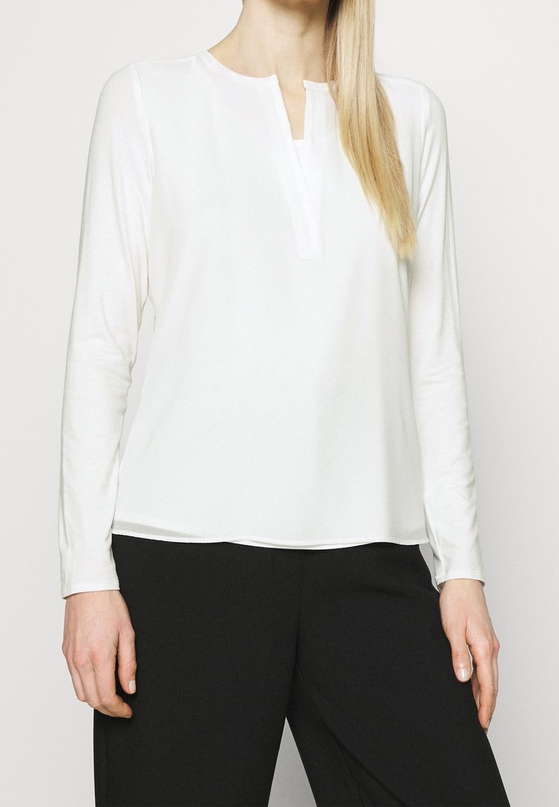Biała bluzka z długim rękawem i delikatnym dekoltem w kształcie litery V, o miękkiej fakturze i prostym wzorze, zestawiona z czarnymi spodniami. Minimalistyczny styl i czyste linie.