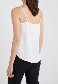 Femme portant un haut sans manches blanc avec des bretelles ajustables et un pantalon noir, vue de dos contre un fond uni.