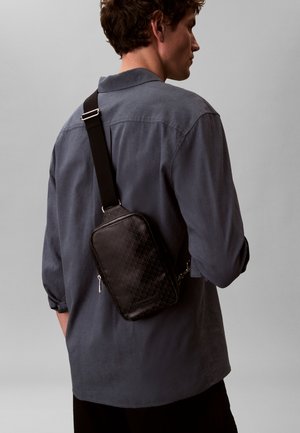 Mann iført mørkegrå langermet skjorte med svart tverrveske med teksturert mønster og sølvglidelås mot ensfarget grå bakgrunn.