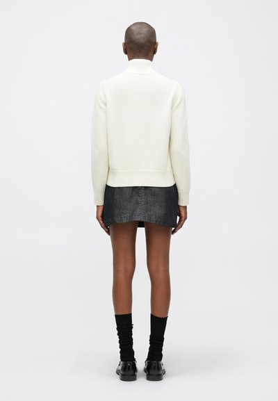 Fehér kötött pulóver magas gallérral, sötét farmer szoknyával párosítva. Fekete zoknik és cipők egészítik ki a megjelenést. Minimalista és lágy textúra.