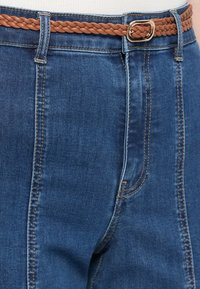 Denim hlače temno modre barve, z izrazitim šivom, pletenim rjavim usnjenim pasom in kvadratno zlatom zaponko.