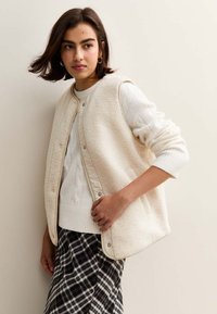 Krämfärgad fleeceväst med tryckknappar över en vit kabelstickad tröja. Modellen bär en svart och vit rutigt kjol, som visar upp lager av texturer.