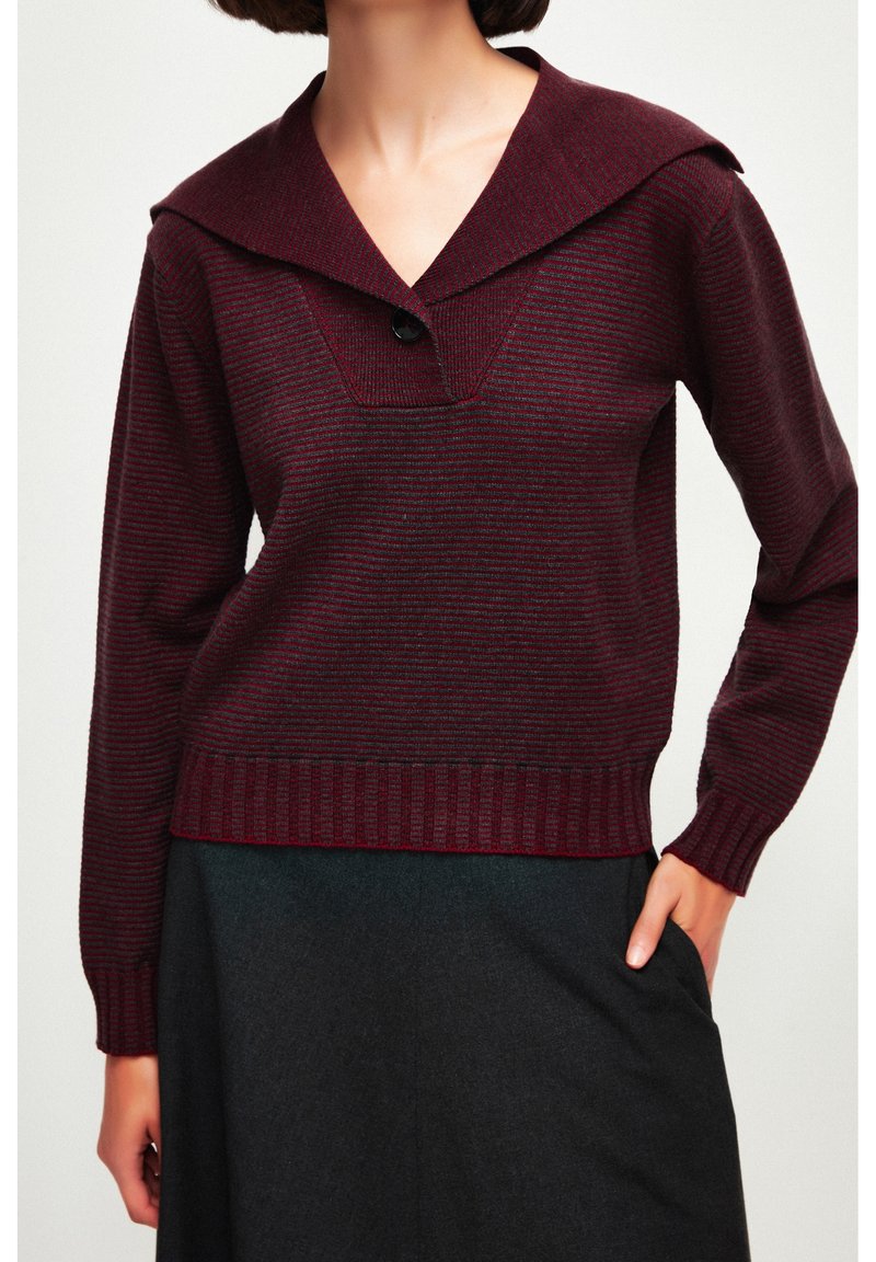 Femme portant un pull en maille rayé rouge foncé avec un col large et un bouton noir, associé à une jupe sombre, la main dans la poche.