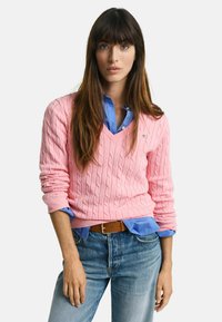 Světle růžový svetr s pleteným vzorem a výstřihem do V, přetáhnutý přes modrou košili s knoflíky. Oblečený s světle modrými džíny a hnědým opaskem.