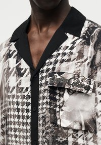 Κοντινό πλάνο πουκάμισου με ασπρόμαυρο μοτίβο καρό, με μαύρο γιακά και τσέπη με καπάκι στο στήθος.
