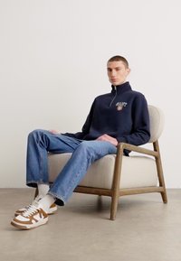 Sweatshirt navy cu fermoar și logo, blugi, și adidași albi cu maro. Modelul este așezat pe un scaun curbat din lemn, pe un podea deschisă la culoare.