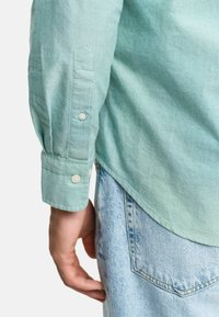 Lichtgroen katoenen overhemd met lange mouwen, knoopmanchetten en een afgeronde onderkant. Draagt over lichtblauwe denim jeans. Zachte textuur met subtiele stikdetails.