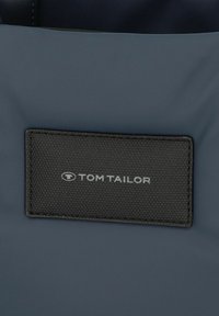 Sac fourre-tout en tissu bleu marine avec une étiquette noire texturée arbore le logo "TOM TAILOR" en lettres argentées. Accents de design minimaliste.