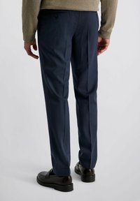 Man in een marineblauwe op maat gemaakte broek en donkerbruine leren instappers, staande met ontspannen handen langs zijn zij op een effen achtergrond.