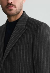 Donkergrijze blazer met een streepjesmotief, met een gesneden revers, een enkele borstzak, gestructureerde schouders en een gestructureerde stof. Close-up weergave.