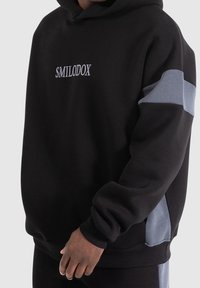 Schwarzer Hoodie mit weißem gesticktem "SMILODOX". Verfügt über graue Seitenpaneele, gerippte Bündchen und eine lockere Passform. Weicher, strukturierter Stoff.