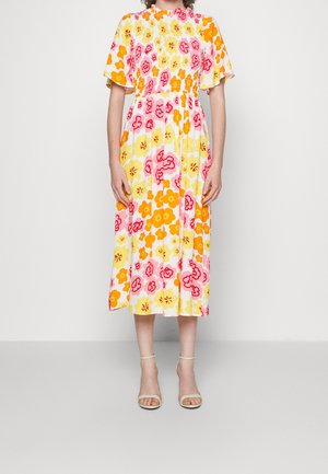 Kvinde iført midikjole med orange, gul og pink blomstermønster, flæseærmer og elastisk talje, kombineret med hvide ankelremssandaler.