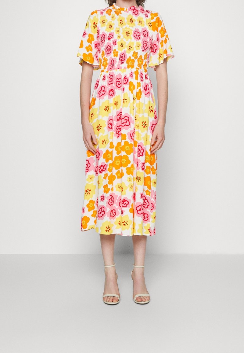 Femme portant une robe midi avec un imprimé floral orange, jaune et rose, des manches volantées et une taille smockée, associée à des talons blancs à bride cheville.