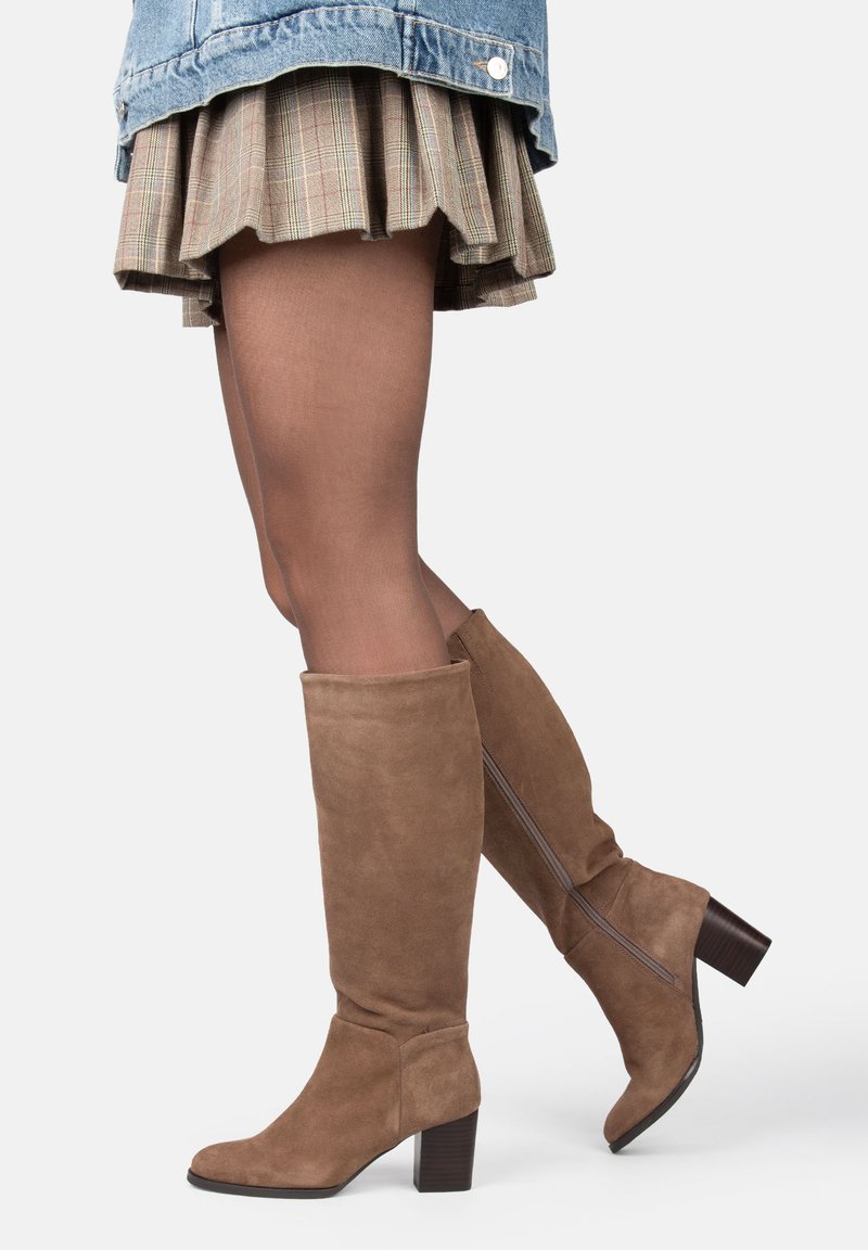 Des jambes portant des bottes en cuir suédé marron jusqu'au genou, des collants transparents marron, une jupe à carreaux et une veste en jean bleue sur un fond blanc.