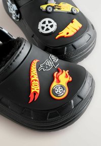 Black rubber clogs with plush lining, featuring colorful 3D rubber decals: flames, racing cars, wheels, and "Hot Wheels" logos.