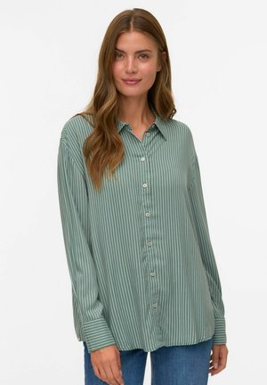 Femme aux longs cheveux châtains clairs portant une chemise à rayures verticales verte et blanche et un jean bleu, se tenant contre un fond blanc.