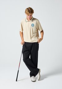 Beige Poloshirt mit Kragen und besticktem Logo, kombiniert mit schwarzen Hosen. Das Model hält einen Golfschläger und trägt weiße Schuhe. Einfaches, sportliches Design.