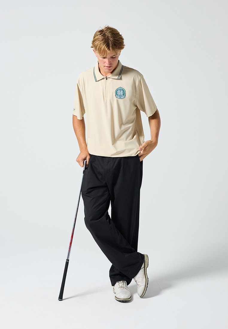 Beige Poloshirt mit Kragen und besticktem Logo, kombiniert mit schwarzen Hosen. Das Model hält einen Golfschläger und trägt weiße Schuhe. Einfaches, sportliches Design.