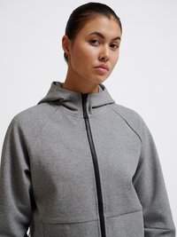 Junge Frau mit dunklen Haaren in einem niedrigen Dutt trägt einen grauen Hoodie mit Reißverschluss und Kapuze vor einem einfarbigen hellen Hintergrund.