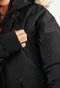 Parka noire avec capuche en fourrure, poche zippée sur la manche, poignets côtelés et un tissu lisse et texturé.