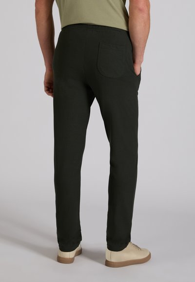 Mężczyzna w czarnych, dopasowanych spodniach i beżowych tenisówkach, stojący tyłem, z rękami w kieszeniach, na jednolitym tle.