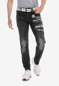 Zwarte denim jeans met verschillende texturen, gestikte patronen, decoratieve studs en meerdere zakken; gedragen met witte sportschoenen.